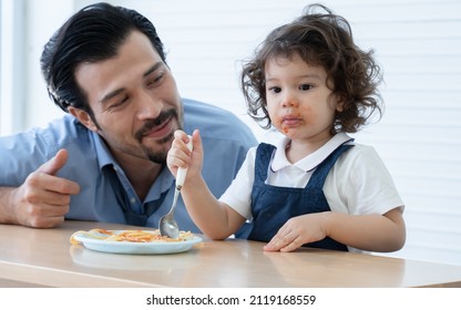 Little Cute Daughter Trying To Eat Spaghetti With Spoon By Herself At Home And Caucasian Young Father Looking And Cheer Her Up. Adorable Kid Girl Enjoy Eating With Face Is Mess Up With Ketchup