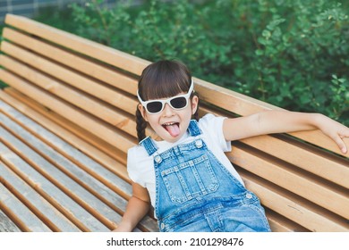 little cute girl 4 years old sits on a bench in summer wearing sunglasses and grimacing looking at the camera, the concept of pampering and childhood.