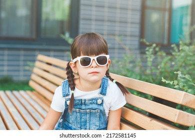 little cute girl 4 years old sits on a bench in summer wearing sunglasses and looking at the camera, the concept of pampering and childhood