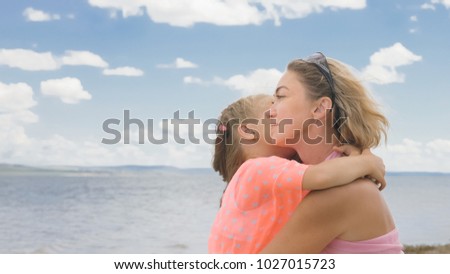 Little cute girl baby and young mother at beach. Mom sits on the shore, and her daughter is coming from behind. The kid hugs and kisses her mother. Mother and her child.