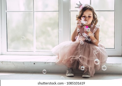 Little cute girl in beautiful dress is sitting near the window at home and blowing soap bubbles.