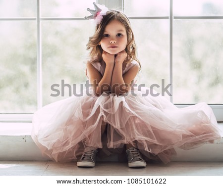 Little cute girl in beautiful dress is sitting near the window at home.