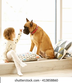 Little Cute Girl With Boxer Dog Sitting On Windowsill At Home