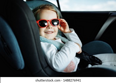 Little Cute Girl In Cap Sitting In The Car In Child Safety Seat And Smiling