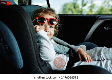 Little Cute Girl In Cap Sitting In The Car In Child Safety Seat And Smiling