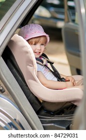 Little Cute Girl In Cap Sitting In The Car In Child Safety Seat And Smiling