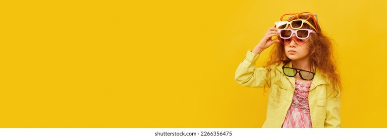 Little cute girl, child with curly hair posing with many colorful sunglasses over yellow studio background. Childhood, emotions, fun, fashion, lifestyle, facial expression. Banner. Copy space for ad