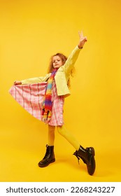 Little cute girl, child with curly hair posing in bright clothes, scarf and boots over yellow studio background. Concept of childhood, emotions, fun, fashion, lifestyle, facial expression