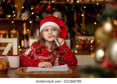 a little cute girl child in a red sweater and a hat in a dark kitchen with a Christmas tree writes a letter to Santa Claus and thinks what to wish for her and waits for the new year or Christmas - Powered by Shutterstock