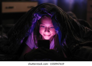 Niña linda a oscuras mira el smartphone bajo una manta en la cama.
