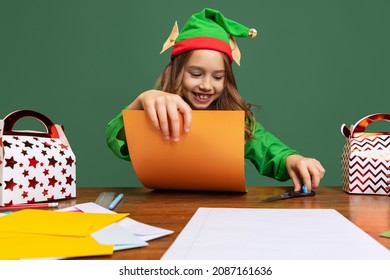 Little cute girl dressed like funny elf sitting at table and making paper Christmas decorations isolated over green background. Merry Christmas and New Year concept. Cute assistant of Santa Claus