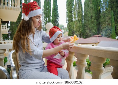 Little Cute Girl And Her Young Mother In Santa Hats In Tropical Country Hotel Balcony. Eating Tangerine. Happy Holidays. Stay Home, Covid, Coronavirus, Merry Christmas. New Year Concept