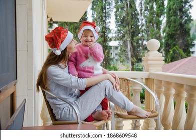 Little Cute Girl And Her Young Mother In Santa Hats In Tropical Country Hotel Balcony. Waiting For Santa. Happy Holidays. Stay Home, Covid, Coronavirus, Merry Christmas. New Year Concept