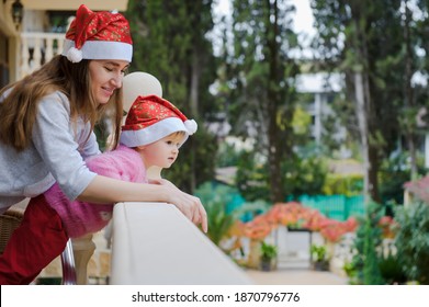 Little Cute Girl And Her Young Mother In Santa Hats In Tropical Country Hotel Balcony. Waiting For Santa. Happy Holidays. Stay Home, Covid, Coronavirus, Merry Christmas. New Year Concept