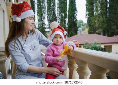 Little Cute Girl And Her Young Mother In Santa Hats In Tropical Country Hotel Balcony. Waiting For Santa. Happy Holidays. Stay Home, Covid, Coronavirus, Merry Christmas. New Year Concept