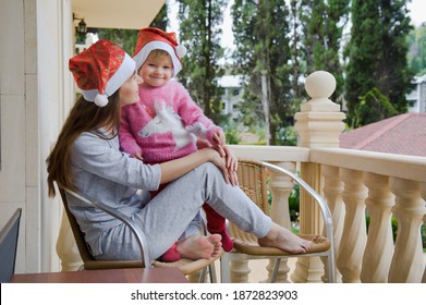 Little Cute Girl And Her Young Mother In Santa Hats In Tropical Country Hotel Balcony. Waiting For Santa. Happy Holidays. Stay Home, Covid, Coronavirus, Merry Christmas. New Year Concept