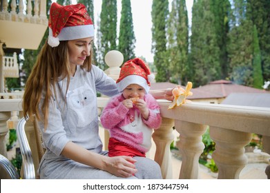 Little Cute Girl And Her Young Mother In Santa Hats In Tropical Country Hotel Balcony. Waiting For Santa. Happy Holidays. Stay Home, Covid, Coronavirus, Merry Christmas. New Year Concept