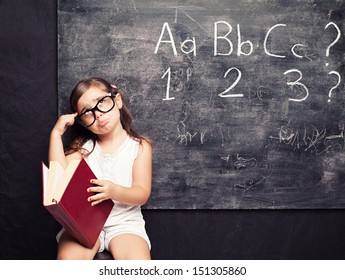 little cute girl holding a red book thinking in front of a blackboard