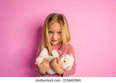 A little cute girl with huge blue eyes and blond hair holds a teddy bear in her arms. The girl cradles a teddy bear. The concept of a happy childhood in pink colors.