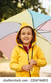 Little Cute Girl Kid Child In A Yellow Raincoat With Multicolored Rainbow Umbrella Laughs And Looks At Camera