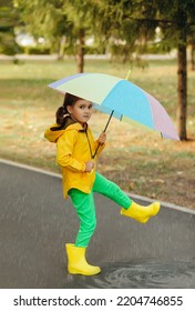 Little Cute Girl Kid Child In A Yellow Raincoat With Multicolored Rainbow Umbrella Walking On A Puddle.