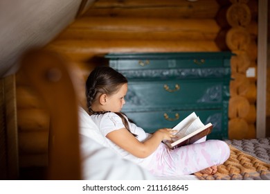 Little, cute girl pictured from side sitting on big bed with book in hands looking at it and reading with chest of drawers in background in wooden house.