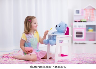 Little cute girl playing with her teddy bear. Child feeding doll in white nursery. Children play with wooden educational toys. Toddler kid in a playroom. Kids feed dolls and cook in toy kitchen.