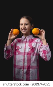 Little Cute Girl Posing With Tangerines In The Studio. Photo Isolated On Black Background With One Light Source. The Child Is Holding A Pair Of Tangerines To His Face.