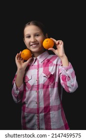 Little Cute Girl Posing With Tangerines In The Studio. Photo Isolated On Black Background With One Light Source. The Child Is Holding A Pair Of Tangerines To His Face.