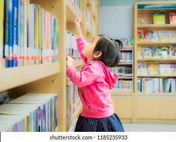 Little Cute Girl Reaching A Book From Bookshelf At The Library. Learning And Education Of Kid.