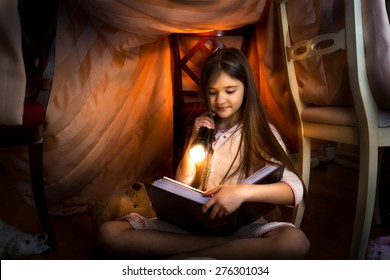 Little cute girl reading book in self-made house with flashlight