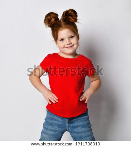 Little cute girl with red hair and freckles holds her hands at her waist and looks to the side. A child in a red T-shirt and jeans poses against a gray wall in the studio.