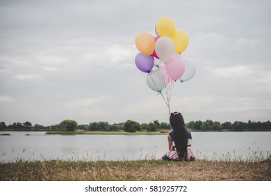 Little cute girl sitting on long green grass outside. Girl holding colorful balloons in hand. 