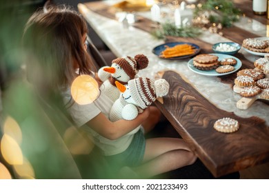 Little cute girl sitting at a table filled with Christmas decor. And he holds two knitted snowmen in his hands. New Year preparation concept