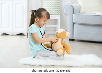 Little cute girl with stethoscope and teddy bear sitting on carpet, on home interior background