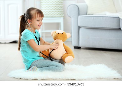 Little cute girl with stethoscope and teddy bear sitting on carpet, on home interior background