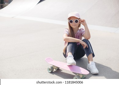little cute girl with sunglasses and cap sitting on a skateboard. photo of cute preteen girl with skateboard outdoors