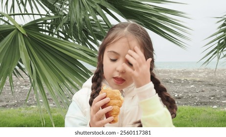 little cute girl with two pigtails on the street near the palm tree on the embankment eating crispy croissant outdoors on street. Close up - Powered by Shutterstock