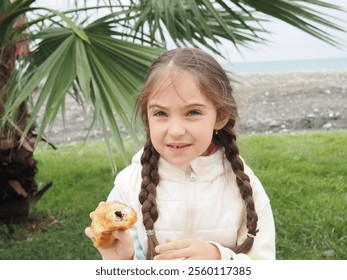 little cute girl with two pigtails on the street near the palm tree on the embankment eating crispy croissant outdoors on street. Close up - Powered by Shutterstock