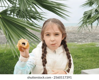 little cute girl with two pigtails on the street near the palm tree on the embankment eating crispy croissant outdoors on street. Close up - Powered by Shutterstock