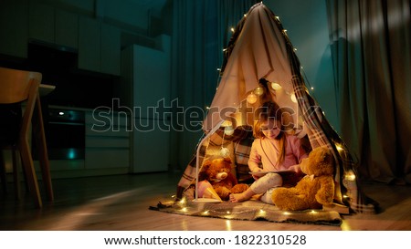 A little cute girl wearing pyjamas sitting on a floor barefoot in a self-made hut made of a plaid, playing games on her tablet, smiling and having teddybears and garlands with her around in a bedroom