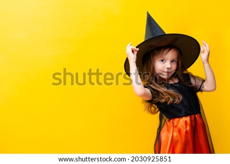 A little cute girl in a witch costume corrects a magic hat. Halloween. Yellow colored background, space for text. High quality photo