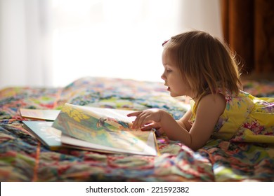 A little cute girl in a yellow dress reading a book lying on the bed