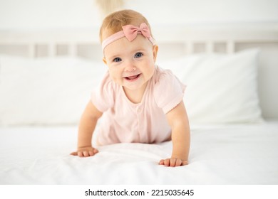 a little cute healthy girl up to a year old in a pink bodysuit made of natural fabric is sitting on a bed on white bed linen in the bedroom, looking at the camera, the baby is at home - Powered by Shutterstock