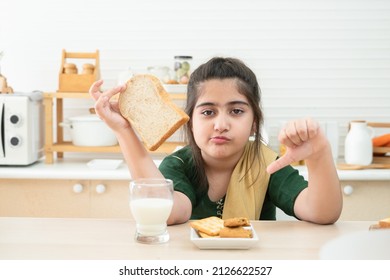 Little Cute Indian Kid Girl Dislike Breakfast Holding Sliced Bread And Thumbs Down While Having Breakfast With A Glass Of Milk, Crackers And Cookies In Kitchen At Home