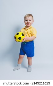 Little Cute Kid Baby Boy 3-4 Years Old, Football Fan In Red T-shirt Holding In Hand Soccer Ball Isolated On Yellow Background. Kids Sport Family Leisure Lifestyle Concept. Copy Space Advertisement