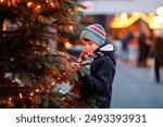 Little cute kid boy having fun on traditional Christmas market during strong snowfall. Happy child enjoying traditional family market in Germany. Schoolboy standing by illuminated xmas tree.