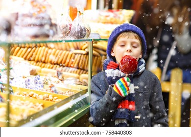 Little Cute Kid Boy Near Sweet Stand With Gingerbread And Nuts. Happy Child Eating On Apple Covered With Red Sugar. Traditional Sweet On German Christmas Market.