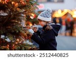 Little cute kid girl having fun on traditional Christmas market during strong snowfall. Happy child enjoying traditional family market in Germany. Schoolgirl standing by illuminated xmas tree.