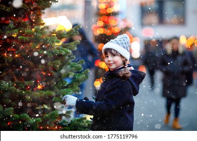 Little Cute Kid Girl Having Fun On Traditional Christmas Market During Strong Snowfall. Happy Child Enjoying Traditional Family Market In Germany. Schoolgirl Standing By Illuminated Xmas Tree.